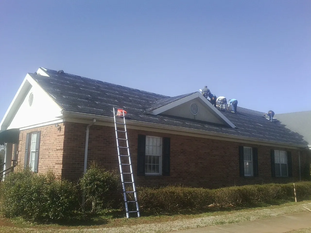 Crew tearing shingles off a brick ranch roof with a ladder set — Emergency Roof Repair work by our crew in North Rose