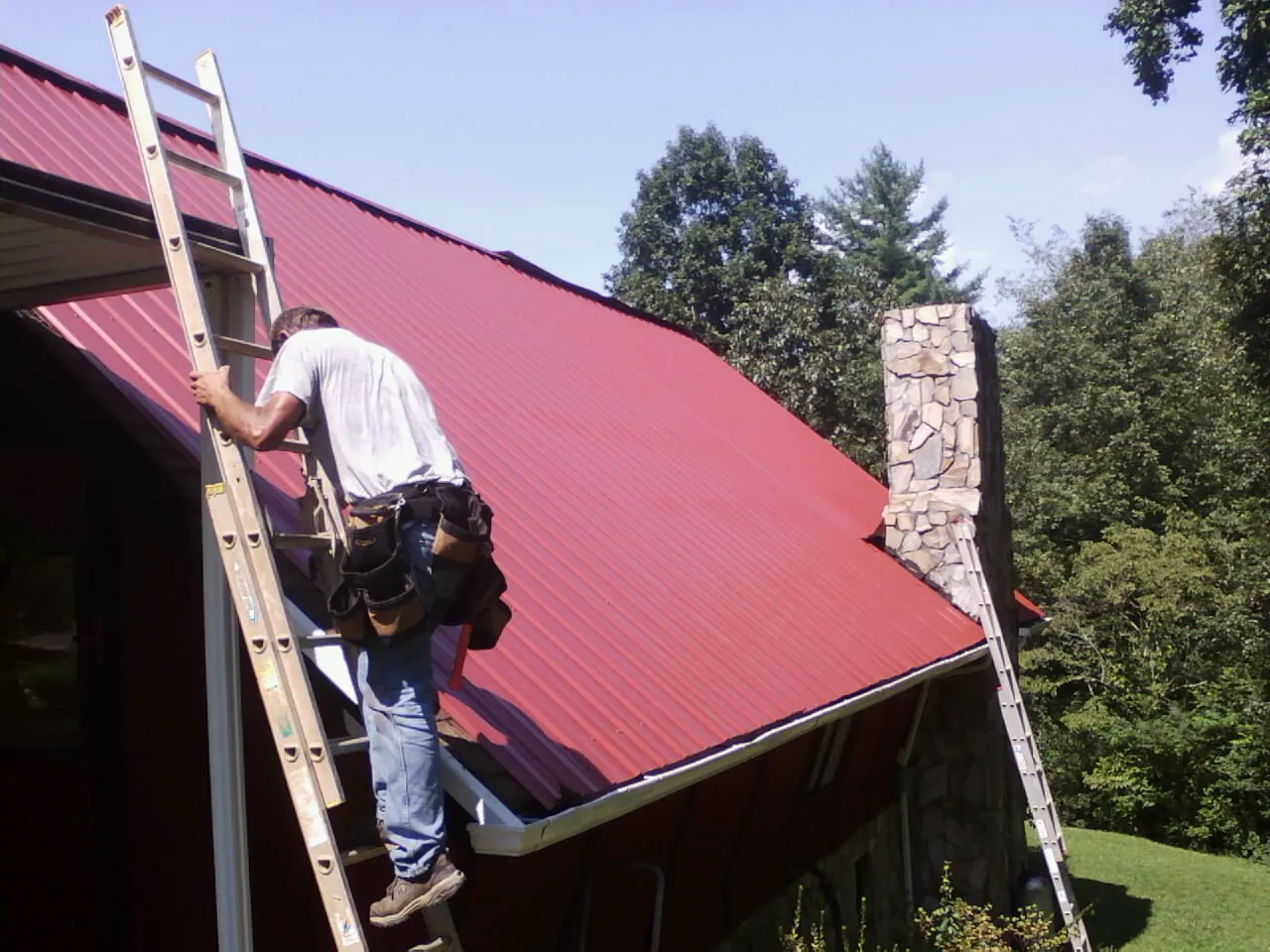 Roofer on a ladder beside a red metal roof and stone chimney — Soffit & Fascia Repair by Roofing Brothers in North Rose